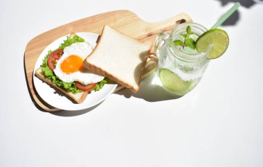 Sandviçli sağlıklı kahvaltı yemeği, kızarmış yumurta, domates dilimi, salata, nane yaprağı, mason kavanozu bardak limonata, ahşap kesim tahtası, beyaz arka plan, güneşli, abur cubur, kilo kaybı, organik, basitlik, vegan, burger, düşük yağ oranı, spor yemeği.