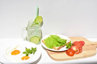 Sandviçli sağlıklı kahvaltı yemeği, kızarmış yumurta, domates dilimi, salata, nane yaprağı, mason kavanozu bardak limonata, ahşap kesim tahtası, beyaz arka plan, güneşli, abur cubur, kilo kaybı, organik, basitlik, vegan, burger, düşük yağ oranı, spor yemeği.