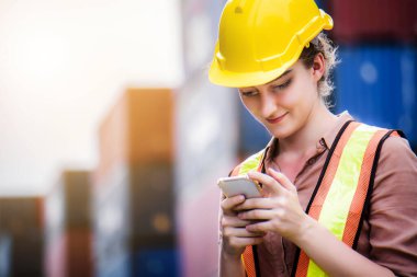 Genç, kendine güvenen beyaz kadın mühendis cep telefonu kullanıyor ve sarı güvenlik kaskı takıyor ve kargo nakliye gemisinden ithalat ve ihracat için konteyner kutusunu kontrol ediyor.