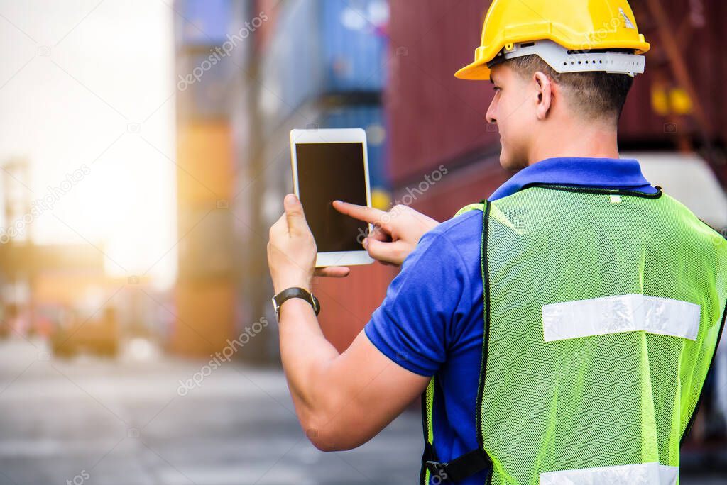 Young confident Caucasian man engineer using digital tablet and wearing yellow safety helmet and check for control loading containers box from Cargo freight ship for import and export, transport 