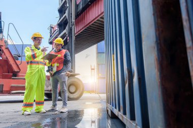 Sarı kasklı mühendis konteyner deposunda forklift kargosunun yanında duruyor ve kargo nakliye gemisinden ithalat ve ihracat için konteyner kutusunu kontrol ediyor. Takım çalışması konsepti 