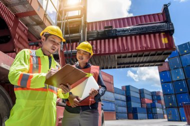 Sarı kasklı mühendis konteyner deposunda forklift kargosunun yanında duruyor ve kargo nakliye gemisinden ithalat ve ihracat için konteyner kutusunu kontrol ediyor. Takım çalışması konsepti 