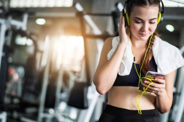 Genç, beyaz bir kadın akıllı telefon ya da cep telefonuna bağlı kulaklıkla müzik dinliyor. Spor salonunda sıkı çalıştıktan sonra rahatlama. 