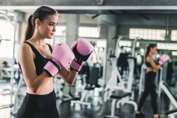 Pretty attractive Caucasian female in pink boxing gloves punching bag during workout in gym.