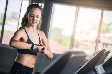 Çekici, beyaz bir kadın koşu bandında koşuyor ve kalp atışlarını spor salonunda kontrol ediyor.. 