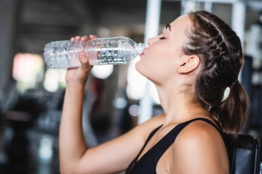 Çekici, beyaz, güzel bir kadın spor salonundaki antrenmandan sonra dinlenip plastik şişeden su içiyor. 