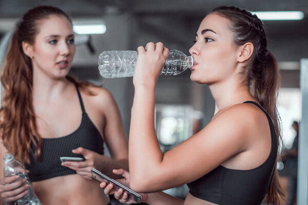 Two young attractive woman talking together and using smart phone or cellphone while relaxation after hard workout at gym 