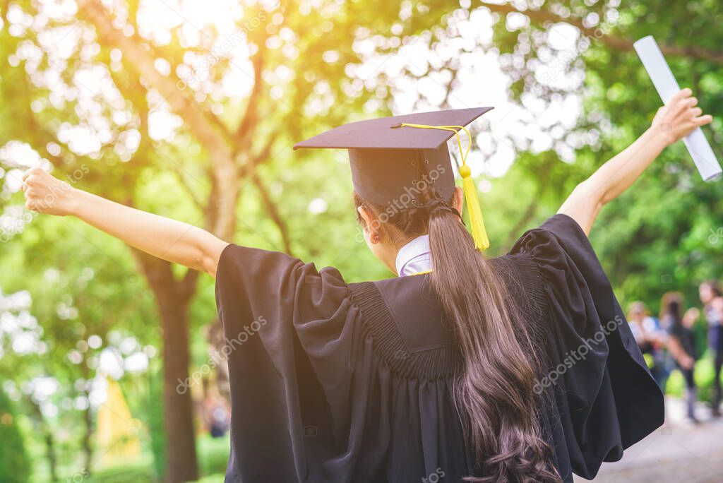 Graduated woman students wearing graduation hat and gown rise her hand with happiness mood with park background 