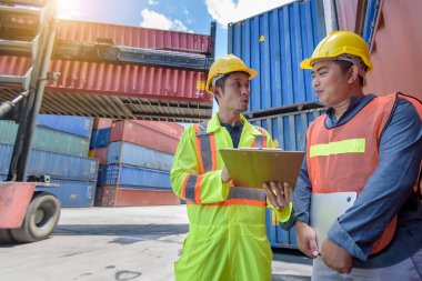 Sarı kasklı mühendis konteyner deposunda forklift kargosunun yanında duruyor ve kargo nakliye gemisinden ithalat ve ihracat için konteyner kutusunu kontrol ediyor. Takım çalışması konsepti 
