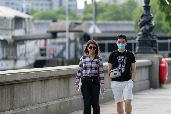Londra, İngiltere 'de Embankment boyunca yürüyen genç Asyalı çift Coronavirus COVID-19' da maske takıyor ve güneşin tadını çıkarıyor.