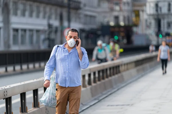 Orta yaşlı Orta Doğulu bir adam Londra, İngiltere 'de Waterloo Köprüsü boyunca yürüyor. Yüz maskesi takıyor ve COVID-19 salgını sırasında cep telefonuyla konuşuyor.
