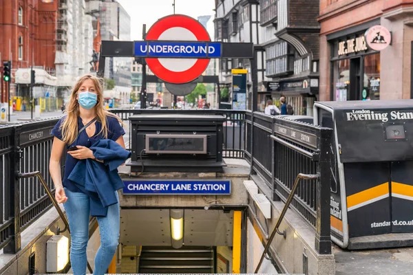 24 Temmuz 2020 'de Chancery Lane Metro İstasyonu' ndan maske takarak çıkan ve COVID-19 Coronavirus salgını sırasında ceketini taşıyan genç sarışın kadın.