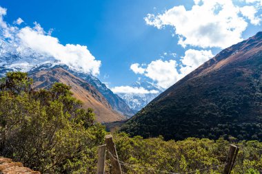 Peru, Cusco 'daki And Vadisi' nde karla kaplı dağlar, yemyeşil bitkiler ve bulutlu mavi gökyüzü. Bu nefes kesici manzara, maceracılar arasında popüler olan Salkantay yürüyüş rotasının bir parçasıdır..