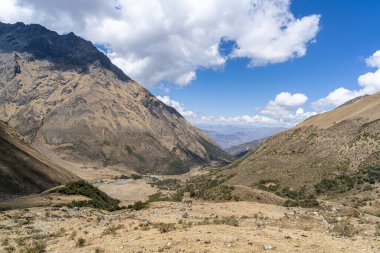 Peru, Cusco 'daki Andean Vadisi' nde kurak dağlar, dağınık bitki örtüsü ve bulutlu mavi gökyüzü. Bu panoramik manzara, yürüyüşçüler ve maceracılar için popüler bir rota olan Salkantay yürüyüşünün bir parçasıdır..