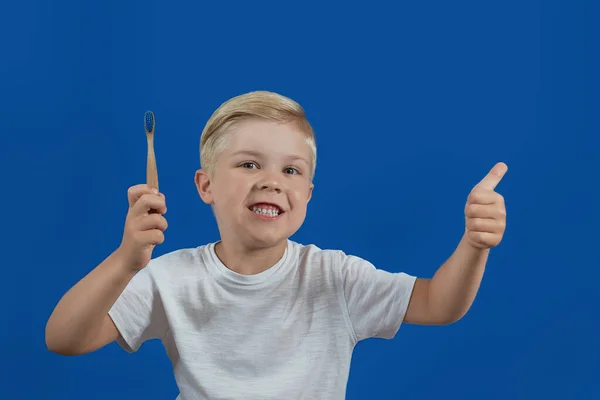 O menino segura uma escova de dentes com pasta e mostra os polegares em um fundo azul — Fotografia de Stock
