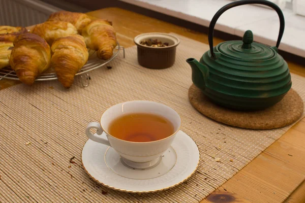 Breakfast or snack, tea with croissant, fine white cup with golden edges with small green kettle for tea, and small brown bowl with tea herbs, on neutral colored textured background.