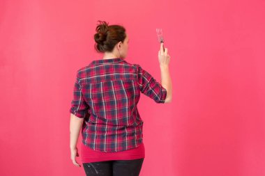 Ekose gömlekli bir kız duvarı pembe boyayla boyuyor, arkadan manzara, pembe arka plan.
