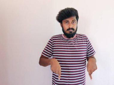 Genç Hintli erkek, doğrudan kameraya bakıyor, çıplak tişört giyiyor, beyaz duvarın karşısında duruyor, ön parmakları aşağıda. Reklam için boşluk kopyala.