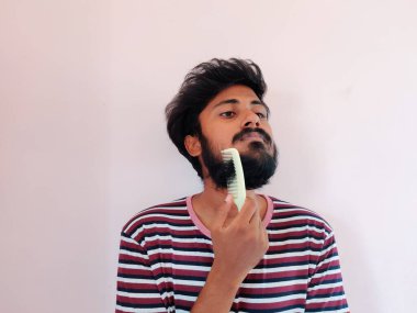 Genç Hintli adam beyaz arka planda sakalını ve bıyığını tarıyor. Adam soyulmuş tişört giymiş. Gün ışığı