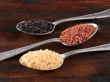 Pişmemiş siyah, kırmızı ve esmer pirinç gümüş kaşıklarda, ahşap bir tahtanın üzerinde 