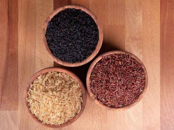 Uncooked black, red and brown rice in olive wood bowls, on a dark rustic wooden boar