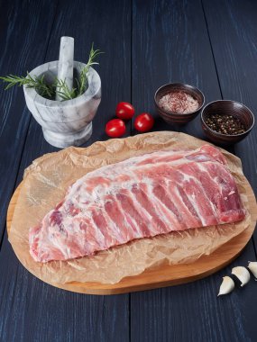 Bambu kesme tahtasındaki buruşuk kağıt üzerinde çiğ domuz kaburgaları, biberiye dalları, domatesler, sarımsak karanfilleri, biber taneleri ve kaba pembe tuz.