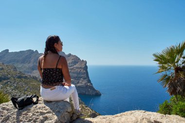 Latin bir kadın, uzun dalgalı esmer saçlı, poz veren, güneş gözlüklü bir bahar gününde İspanya 'nın Balear Adaları' ndaki Mallorca Cap de Formentor 'un uçurumunda oturuyor.,