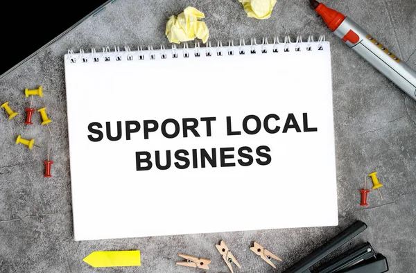 Support local businesses text on a white notebook with pins, marker and stapler on a concrete table