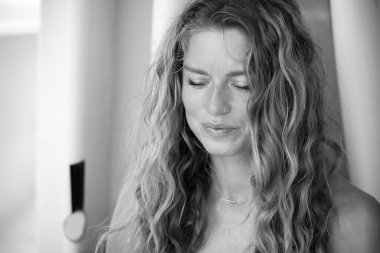 Siyah beyaz bikinili genç bir kadının portresi. Arka planda sörf tahtaları var. Gülümsüyor.