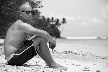 Güneş gözlüklü İskandinav kel adamın tropik plajda oturan siyah ve beyaz portresi