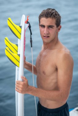 Beyaz dişli genç adamın portresi gülümsüyor, elinde sarı yüzgeçli beyaz sörf tahtası ve mavi okyanus ve arka planda tropikal ada tutan sörfçü.