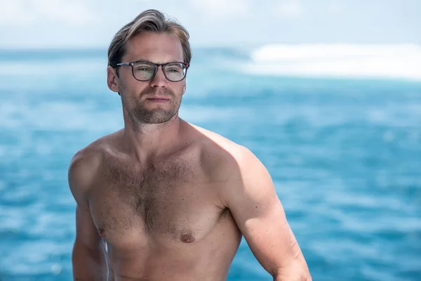 Portrait of handsome shirtless man, surfer in stylish glasses on surfsafari boat and blue ocean with waves on background