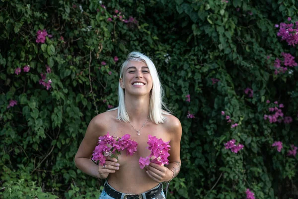Portrait of young blonde woman with pink flowers cover breast and green bush with pink flowers on background