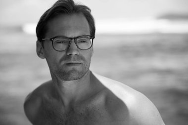 Yakışıklı üstsüz bir adamın portresi, sörf teknesinde gözlüklü bir sörfçü ve arkasında dalgalar olan mavi bir okyanus.