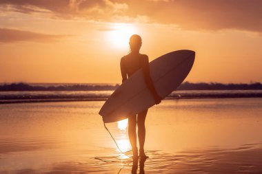 Bali 'de gün batımında sörf tahtasıyla sahilde güzel vücutlu sörfçü kızın portresi.