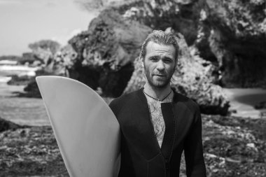 Siyah dalgıç kıyafetli yakışıklı sörfçünün siyah beyaz portresi, beyaz sörf tahtası ve mavi okyanus, uçurum, arka planda kayalar.