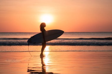 Bali 'de günbatımında sörf tahtasıyla sahilde güzel vücutlu bir kadın sörfçü portresi