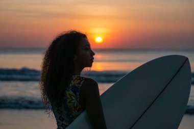 Bali 'de günbatımında sörf tahtasıyla sahilde güzel vücutlu bir kadın sörfçü portresi