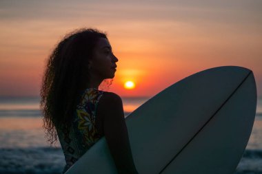 Bali 'de günbatımında sörf tahtasıyla sahilde güzel vücutlu bir kadın sörfçü portresi