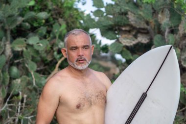 Beyaz sörf tahtası ve Bali arka planında yeşil kaktüs tutan yakışıklı sörfçü portresi.