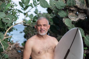 Beyaz sörf tahtası ve Bali arka planında yeşil kaktüs tutan yakışıklı sörfçü portresi.