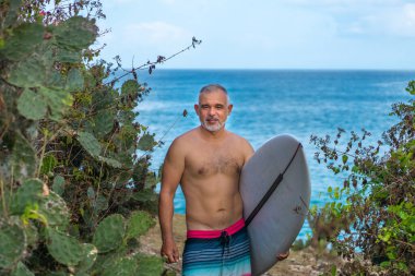 Beyaz sörf tahtası ve Bali arka planında yeşil kaktüs tutan yakışıklı sörfçü portresi.