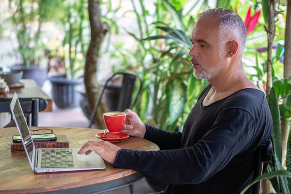 Mature man freelancer working with laptop and phone holding red cup of coffee in Bali