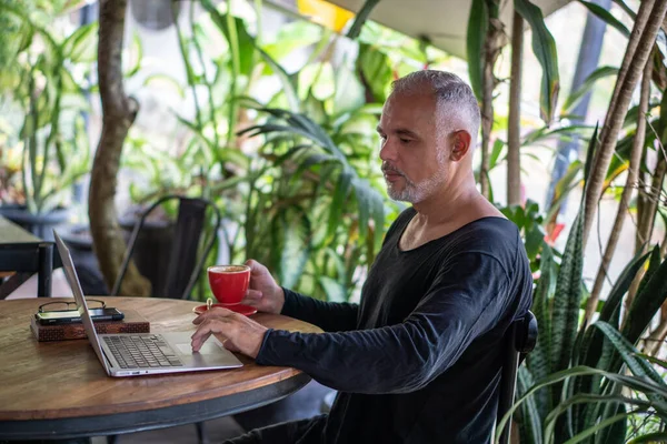 Mature man freelancer working with laptop and phone holding red cup of coffee in Bali