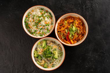 Üç Asya mezesi yuvarlak kase pirinç, yumurtalı erişte, cam erişte ve sebze. Pan-Asya ve Asya yemekleri ve yemekler şık, siyah beton mutfak masasında..