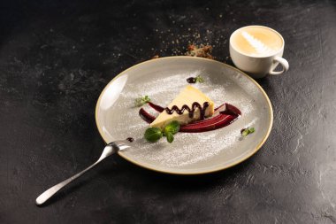Kahveli klasik tatlı - böğürtlen soslu cheesecake, nane yapraklarıyla süslenmiş ve seramik tabakta toz şeker ve siyah taş mutfak masasında bir fincan kapuçino..