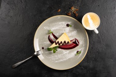 Kahveli klasik tatlı - böğürtlen soslu cheesecake, nane yapraklarıyla süslenmiş ve seramik tabakta toz şeker ve siyah taş mutfak masasında bir fincan kapuçino..
