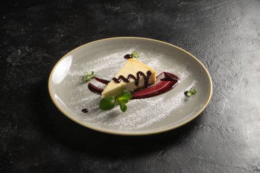 Klasik tatlı - böğürtlen soslu cheesecake, siyah taş mutfak masasında yuvarlak bir seramik tabakta nane yaprağı ve pudra şekerle süslenmiş