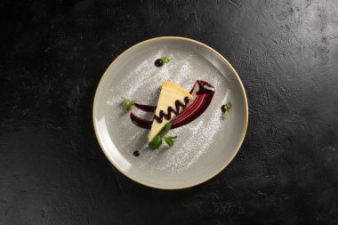 Klasik tatlı - böğürtlen soslu cheesecake, siyah taş mutfak masasında yuvarlak bir seramik tabakta nane yaprağı ve pudra şekerle süslenmiş