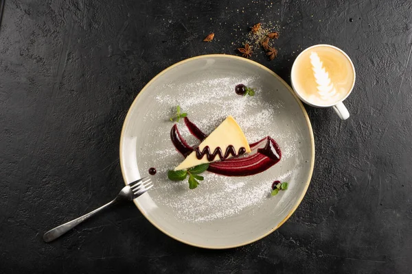 Kahveli klasik tatlı - böğürtlen soslu cheesecake, nane yapraklarıyla süslenmiş ve seramik tabakta toz şeker ve siyah taş mutfak masasında bir fincan kapuçino..